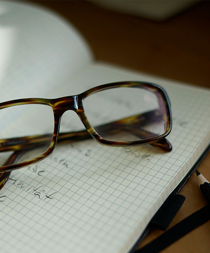 Brille liegt auf kariertem Notizblock mit handschriftlichen Notizen und Stift auf Schreibtisch