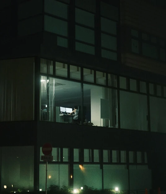 Beleuchtetes Büro bei Nacht mit Person, die allein am Schreibtisch arbeitet hinter großer Glasfassade